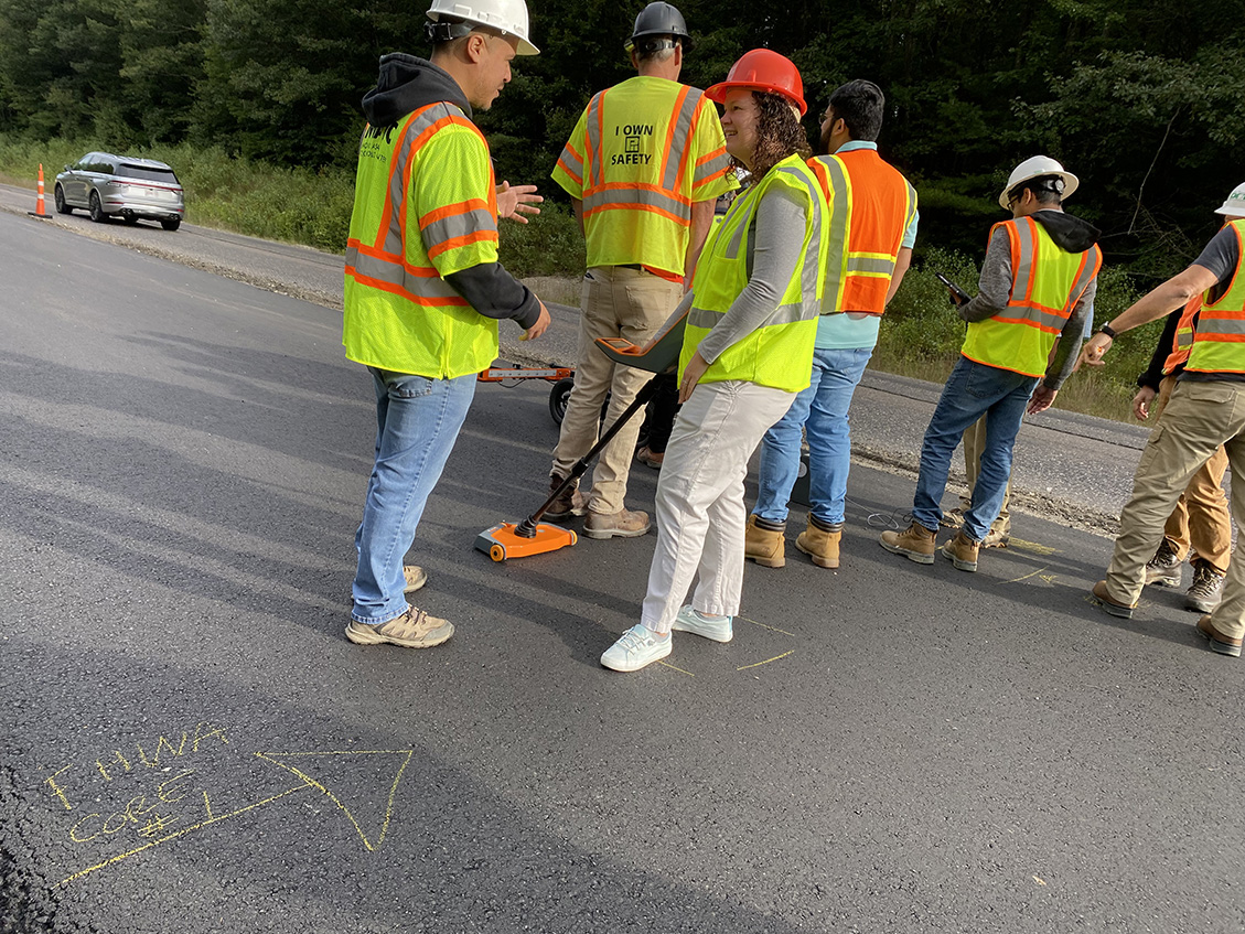 Group of people outdoors on an active asphalt paving job wearing personal protection equipment and learning to use field technologies for post-construction.